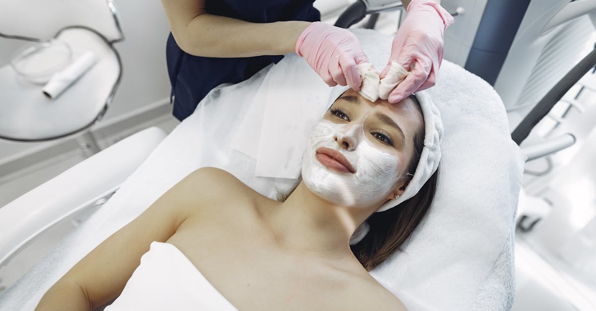 Unrecognizable crop female beautician in medical coat and latex gloves applying facial treatment of young woman resting on couch in modern spa studio