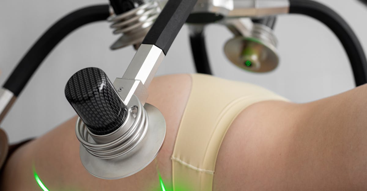 Side view of crop anonymous female in underwear lying and doing non invasive laser body treatment against light wall in clinic